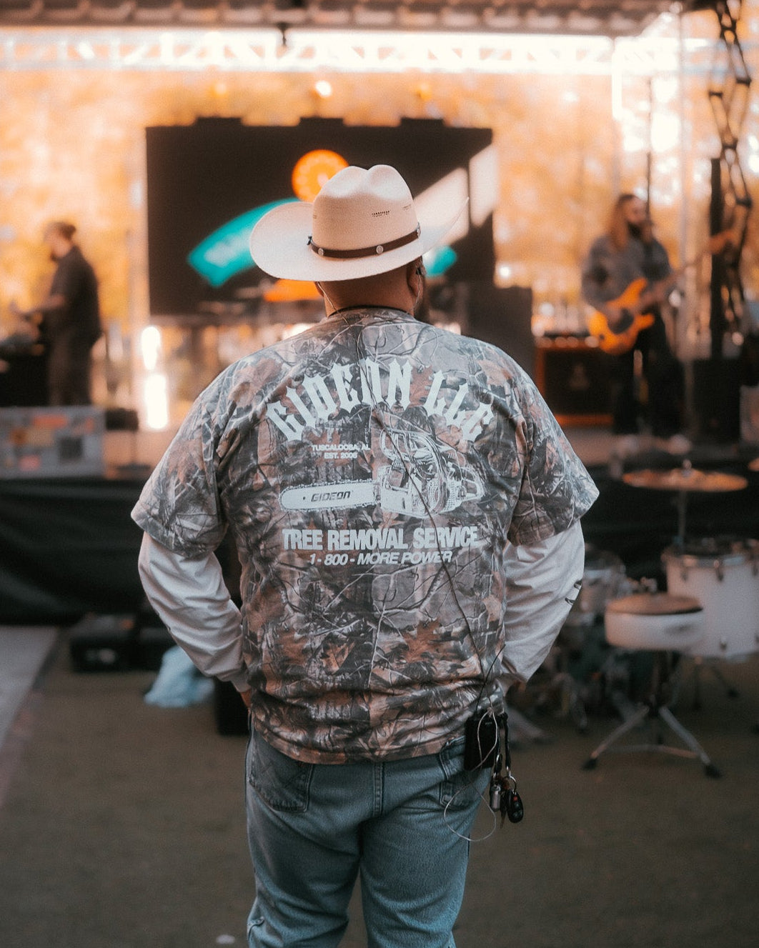 DANIEL MCWHORTER OF GIDEON. WEARING LONG-SLEEVE CAMO TEE WITH WHITE LONG SLEEVES. SHOWING BACK PRINT OF GIDEON LLC TREE REMOVAL SERVICE.